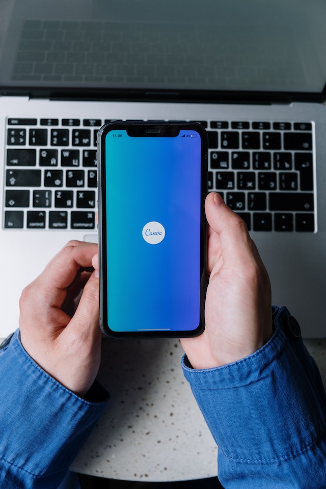 A hand holding a phone with the Canva logo above a Macbook.