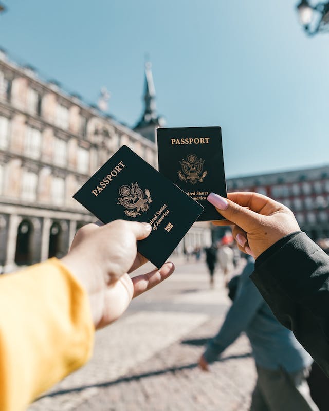 Two people hold up their passports for a photo in a town square.