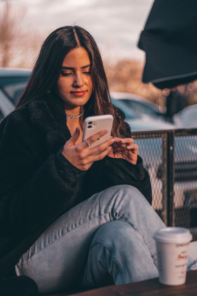 Kobieta uśmiecha się do swojego telefonu, siedząc przed kawą.