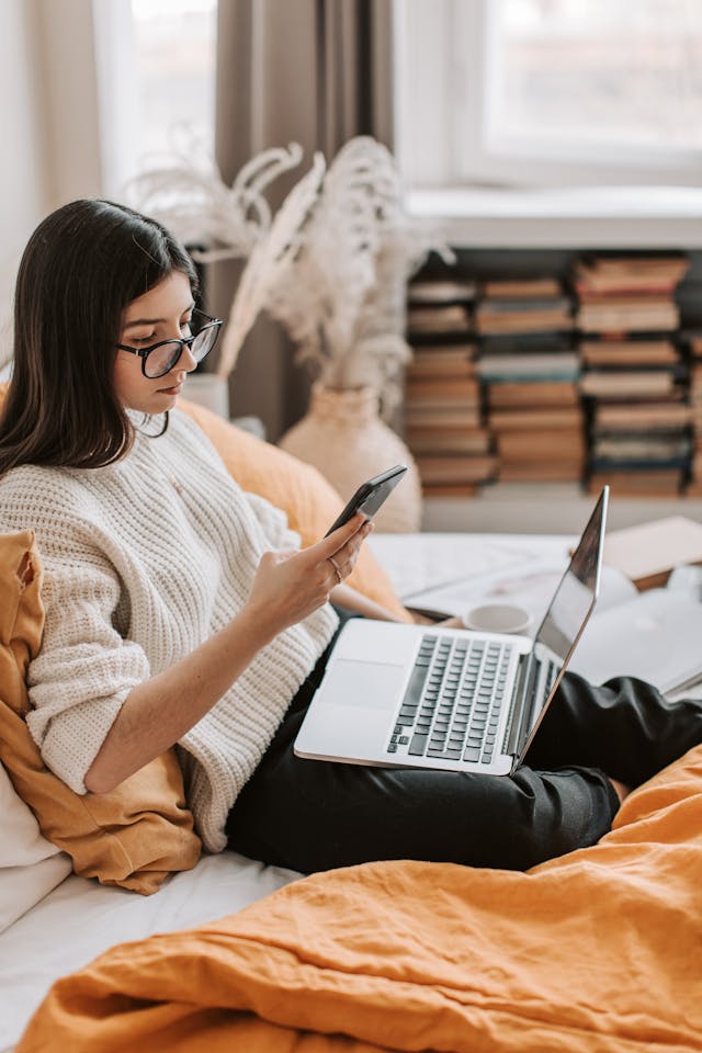 Una donna scorre il suo telefono e usa un computer portatile a letto.