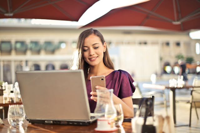 Kobieta siedzi przed laptopem i uśmiecha się do swojego telefonu podczas posiłku w restauracji.