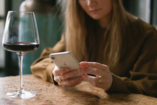 A woman taps on her phone beside a glass of wine.